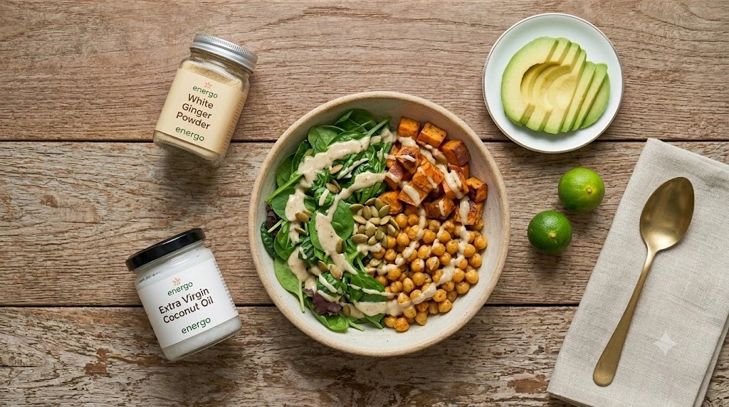 A top-down view of a healthy roasted sweet potato and chickpea nourish bowl with spinach and creamy tahini drizzle, styled on a wooden table next to individual jars of Energo Extra Virgin Coconut Oil and White Ginger Powder.